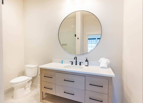 A white bathroom with a round mirror and a white toilet at Westgate Apartment Homes, Pendleton, OR