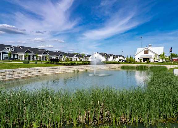 A white building with a flag on top stands behind a pond with a fountain in the middle.