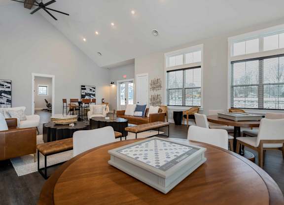 A large wooden table with a checkerboard in the middle is in the foreground of a spacious room with multiple seating areas and large windows.