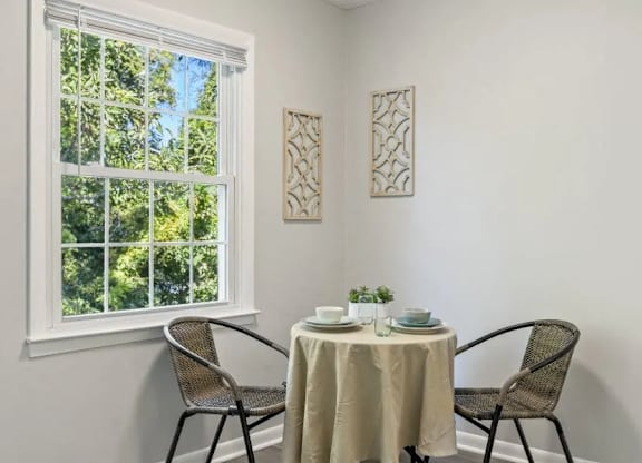 A table with a white tablecloth and two chairs is set up in front of a window at The Preserve at Pine Valley Apartments, North Carolina 28412