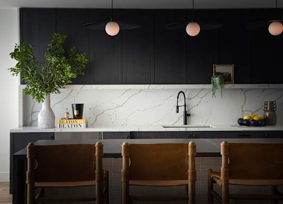 A kitchen with black cabinets and a marble backsplash with three chairs in front.