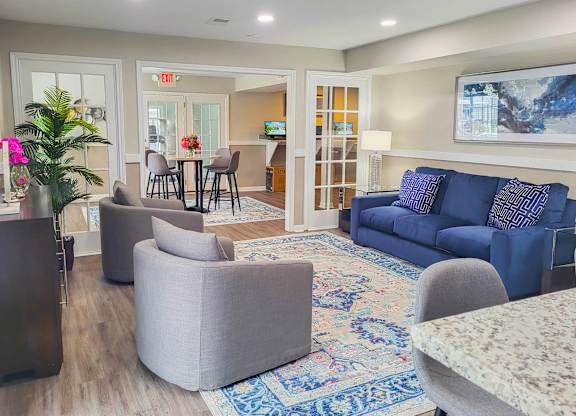 A living room with a grey sofa, a grey armchair, a grey ottoman, a wooden side table, a glass table with chairs, a rug, and a painting on the wall.