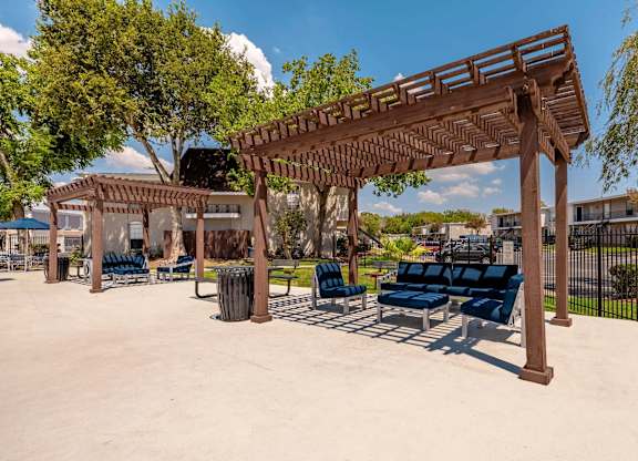 A wooden pergola with blue cushioned seating is in the foreground of a sunny park.