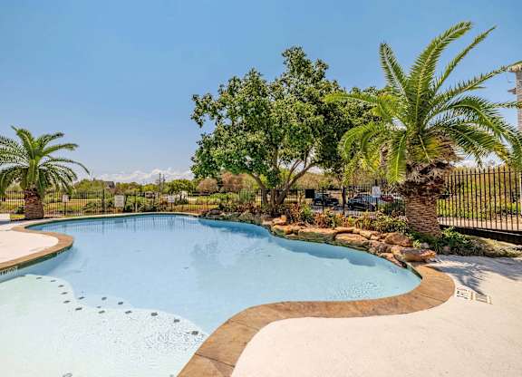 A large outdoor swimming pool surrounded by palm trees and a fence.