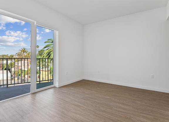 A room with a sliding glass door leading to a balcony with a view of a fence and trees.