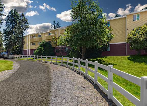 A white fence runs along a gravel road in front of apartment buildings.