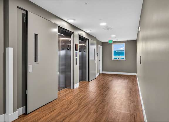 A long hallway with wood floors and a row of elevators.