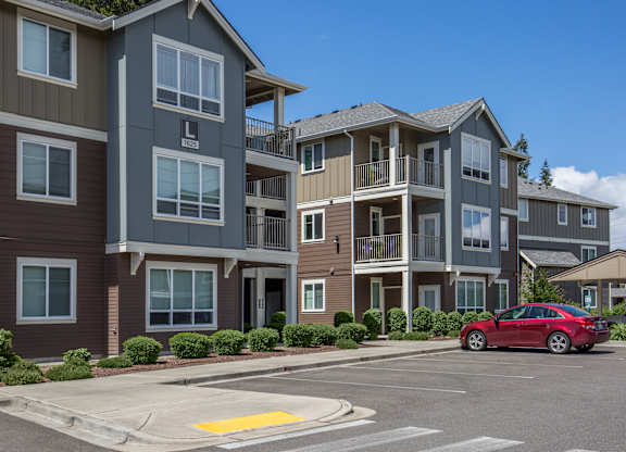 A red car is parked in a parking lot in front of apartment buildings.