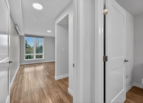 A hallway with white walls and wooden floors leading to a bright window.