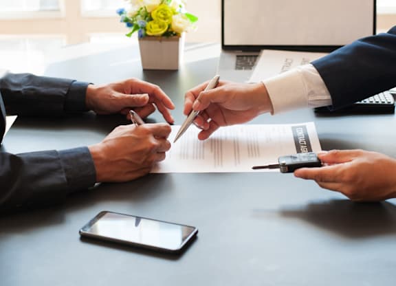 Two people are signing a document with a pen while holding a smartphone and a calculator on the table.