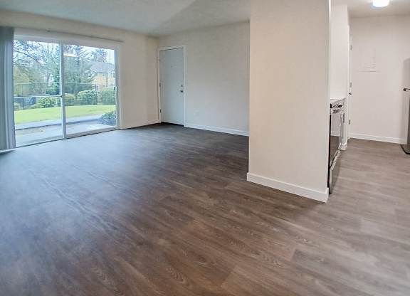 A spacious room with wooden flooring and a view of a green outdoor area through the windows.
