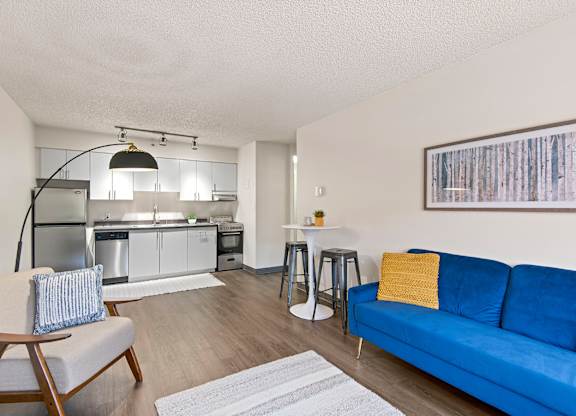 Tacoma, WA, Apartments Near St. Joseph Medical Center - The Orion - Open Living Room With Blue Sofa, Wood-Style Flooring, Floor Lamp, Armchair, Stools, and Wall Art.