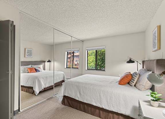 Apartments Near St. Joseph Medical Center, Tacoma, WA - The Orion - Bright Bedroom With Mirrored Closet Doors, Neutral Carpet, White Bedding, Bedside Lamps, and Window.