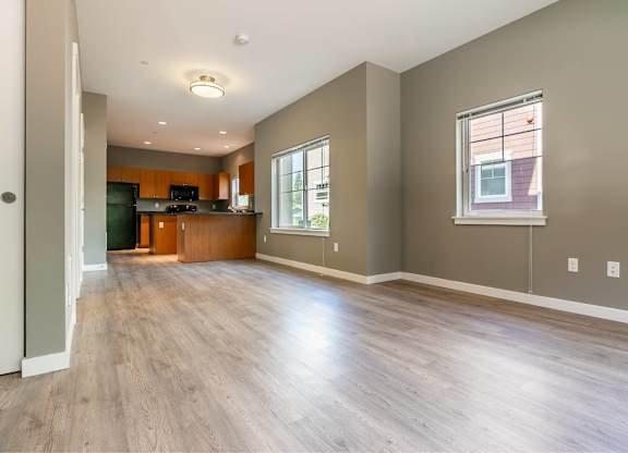A spacious living room with wood flooring and a kitchen area in the background.