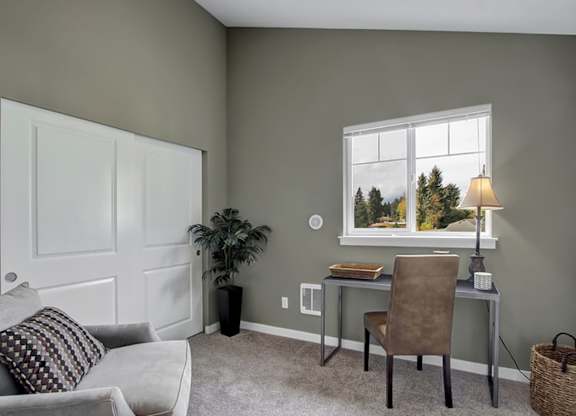 A room with a grey couch, a brown chair, a desk with a lamp and a window showing trees outside.