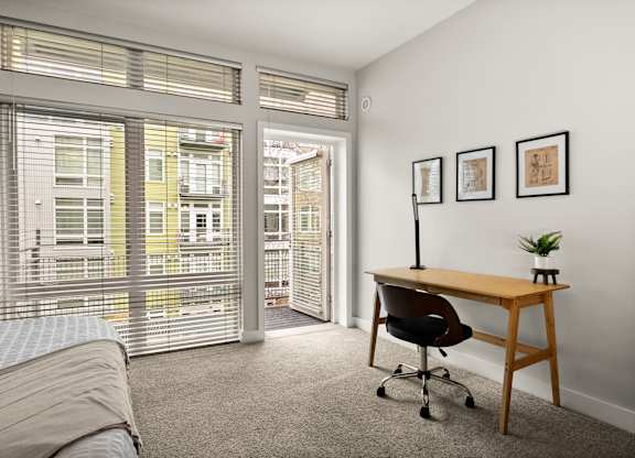 A room with a bed, desk, and chair with a window overlooking a balcony.
