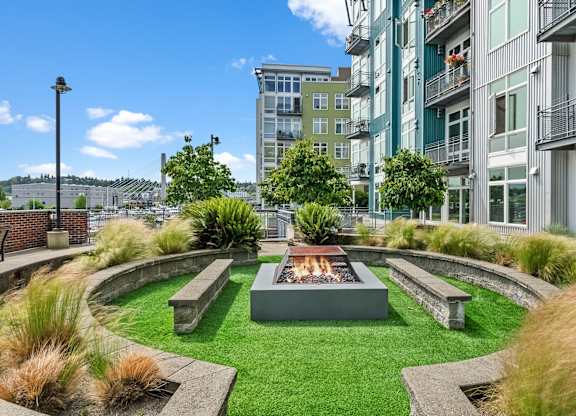 A modern outdoor fire pit is surrounded by a grassy area and concrete benches.