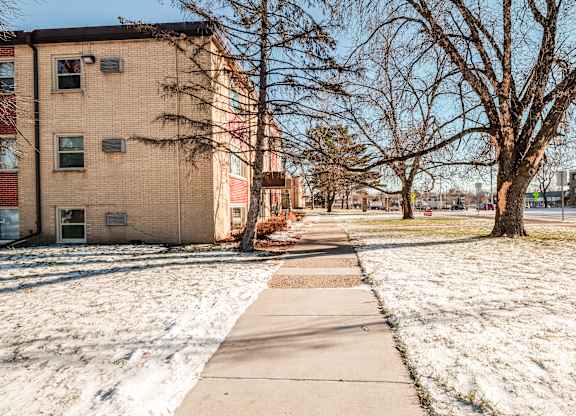 A snow-covered ground with a sidewalk and a tree in front of a brick building.