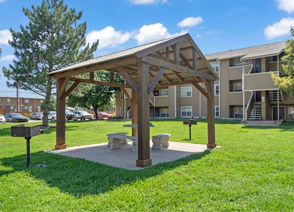 A wooden pavilion with a concrete base is surrounded by a grassy area.