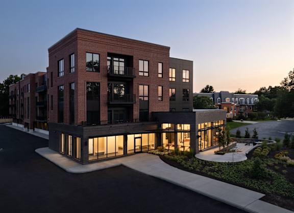 A modern multi-story building with a brick facade and large windows is illuminated from within, showcasing its interior lighting.