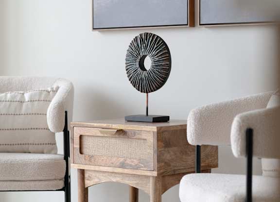 A room with a white chair, a wooden table with a fan on it, and a painting on the wall.