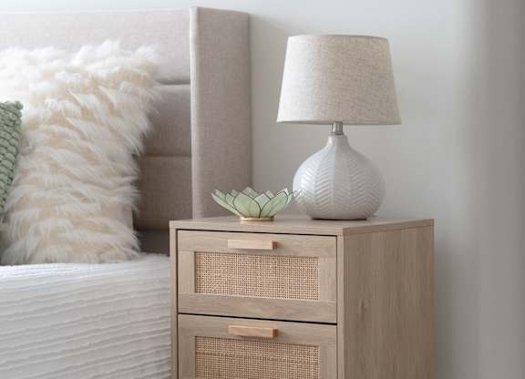 A white lamp with a green plant on top of a wooden nightstand.