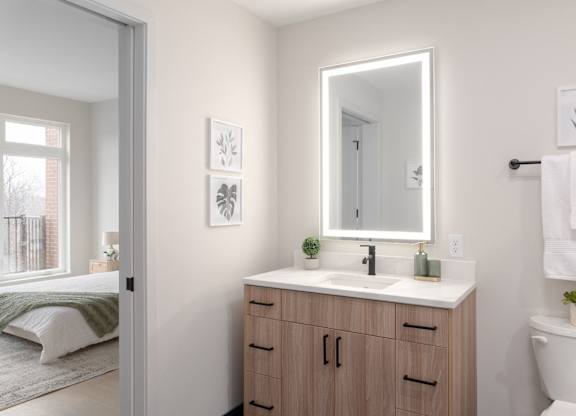 A bathroom with a white toilet, a sink with a mirror above it, and a bedroom with a bed and a window.