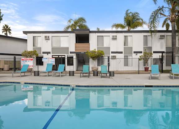 A swimming pool in front of a building with a fence and chairs around it.