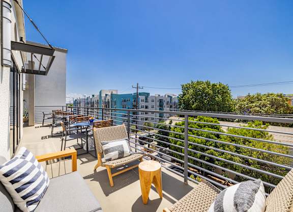 A balcony with a grey couch, wooden chairs and a table with a striped pillow.