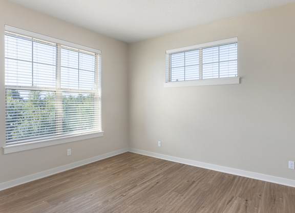 A room with wooden flooring and two windows with blinds.