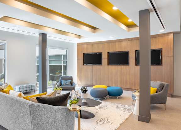 A modern living room with a grey sofa, yellow and blue cushions, a wooden cabinet, and a large window.