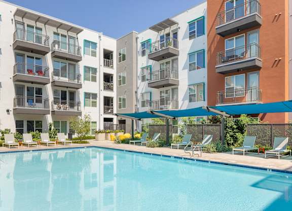A swimming pool is surrounded by lounge chairs and umbrellas in front of apartment buildings.