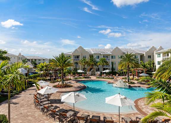 A large swimming pool surrounded by lounge chairs and umbrellas with a resort in the background.