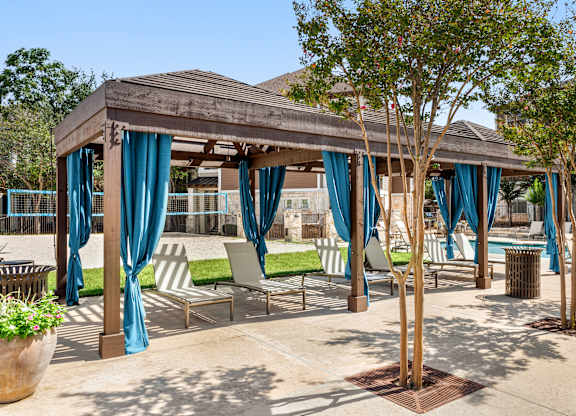A gazebo with blue curtains is surrounded by chairs and trees.