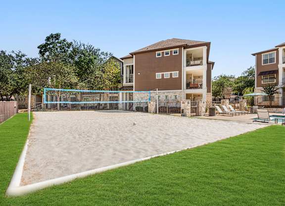 A sand volleyball court is located in a grassy area in front of apartment buildings.