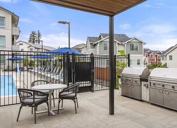 A patio with a table and chairs is surrounded by a pool and houses.
