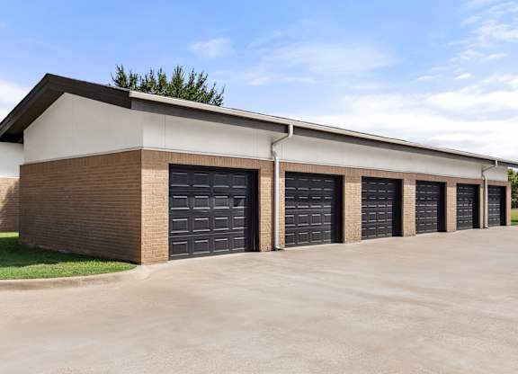 A building with a brown brick facade and a white roof has six black garage doors.