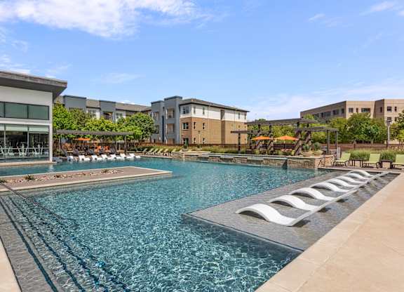 A large swimming pool with lounge chairs is surrounded by buildings.