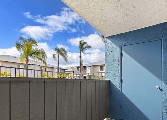 A blue door is on the right side of a wall with a balcony in the background.