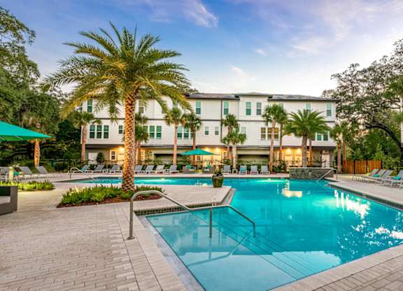 A swimming pool surrounded by lounge chairs and palm trees in front of a hotel.
