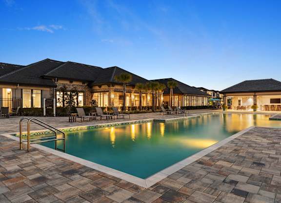 A large outdoor swimming pool surrounded by a stone patio and a building with a black roof.