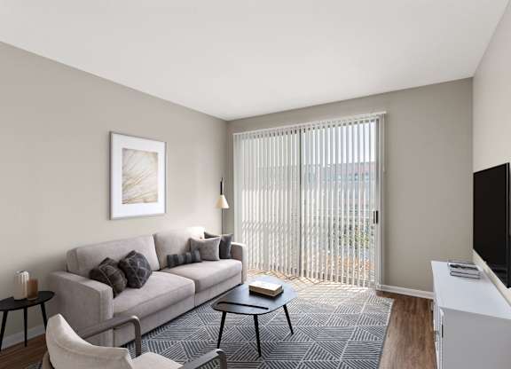 A living room with a grey couch, a black chair, a small table, a television, and a window with blinds.