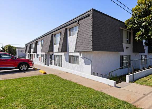 A red car is parked in front of a white building with a black roof.