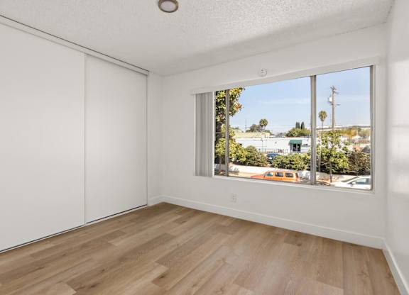 A room with white walls and wooden flooring with a window showing a view of a street with cars and trees.