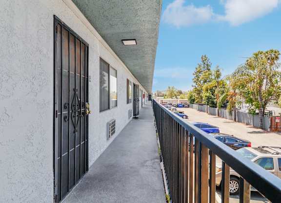 A long, narrow balcony with a metal railing and a white wall with a black gate.