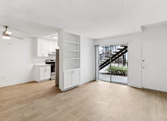 A room with a wooden floor and white walls, with a kitchen area and a staircase visible through the glass door.