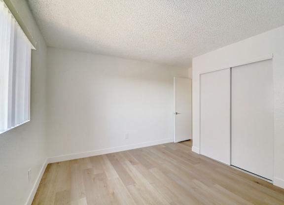 A room with white walls and wooden flooring with a window on the left.