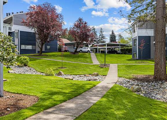 A pathway leads through a grassy area with a tree and a building in the background.