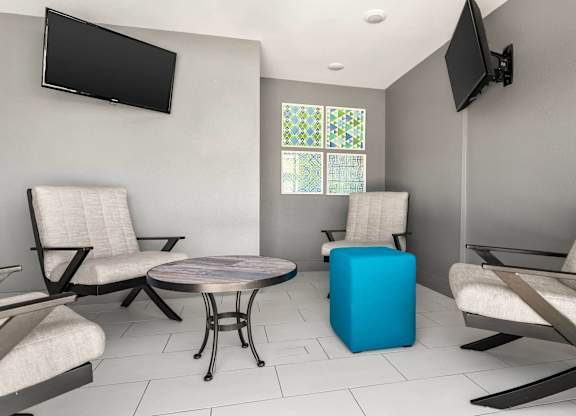 A living room with two chairs, a table, and two TVs mounted on the wall.