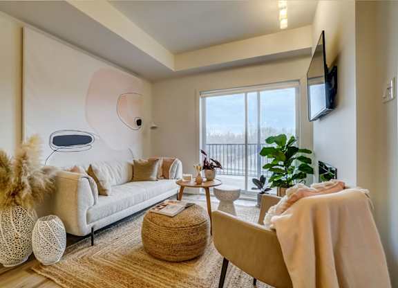 A living room with a white couch, a brown ottoman, a white chair, a brown wicker chair, a white wall with a TV, a window with a view of the outside, and a plant at Champlain Village Urbain Apartments 